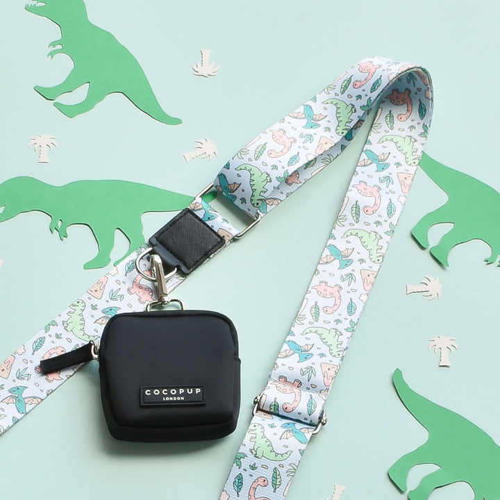 A small black pouch labeled "Cocopup London" is attached to the Bag Strap - Dinopaws, which boasts a pastel-colored dinosaur print on a mint green background with cut-out green dinosaur shapes and small palm trees.