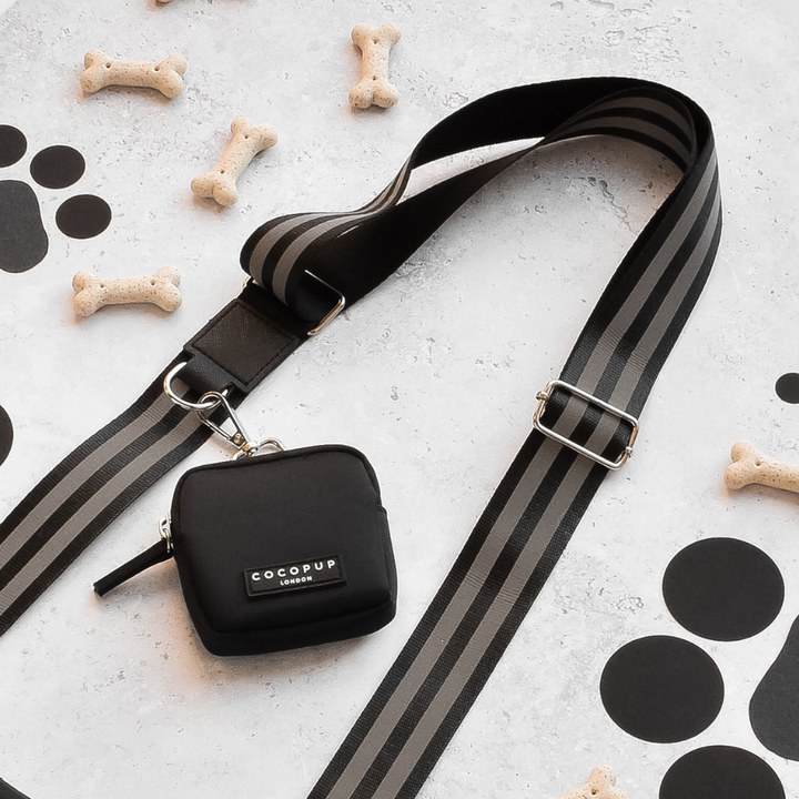 A black and gray striped reflective bag strap, featuring a small black pouch with "Cocopup London" written on it, lies on a white surface. Perfect for walking your dog during late night walks, the strap is surrounded by several small dog bone-shaped biscuits and scattered black paper circles.