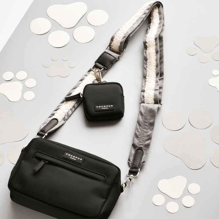 A black crossbody dog walking bag accompanied by a matching small pouch, both emblazoned with the "Cocopup London" logo. The bag's strap is a "Bag Strap - Grey Aviator" with a cozy fleece lining. These items are displayed on a gray surface decorated with light gray paw print cutouts.