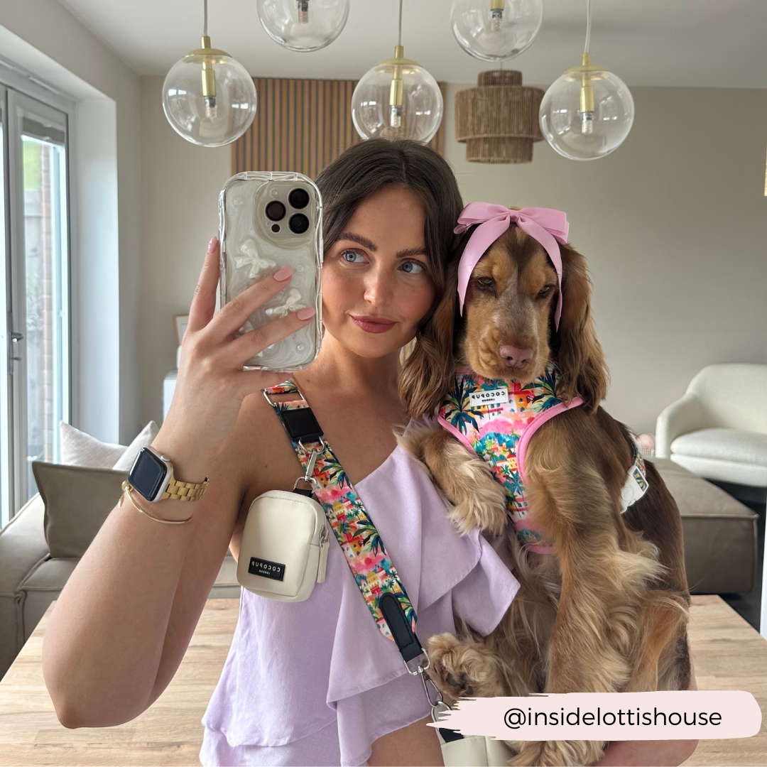 A woman with long brown hair takes a mirror selfie while holding a brown dog that is sporting a pink bow and floral harness. She wears a light purple top and carries a white crossbody bag accented with the Portofino Pup Bag Strap by Cocopup London. The modern dining area in the background features glass pendant lights. Instagram: @insidelottishouse