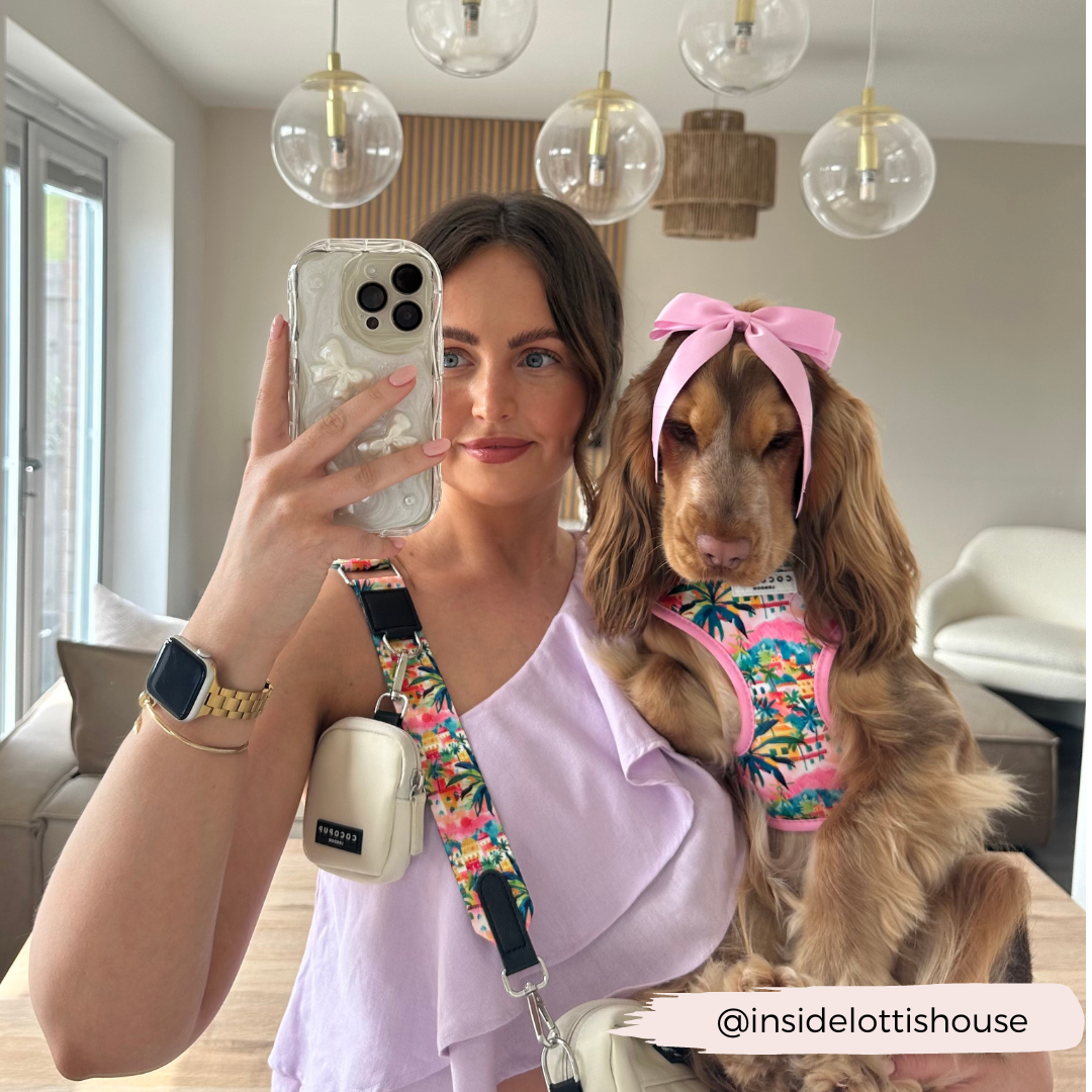 A woman in a lavender top takes a mirror selfie while holding a brown dog dressed in a colorful outfit with a pink bow and accessorized with the stylish Bag Strap - Portofino Pup by Cocopup London. They are in a modern room with hanging lights and a wooden table. The Instagram handle "@insidelottishouse" is overlaid in the corner.