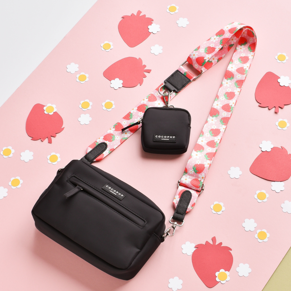 A black crossbody bag with a detachable "Pupberry Fields" strap and a matching small black pouch is laid out on a pink and red background decorated with strawberry and white flower cutouts. Perfect for stylish dog walks, the strap and pouch both feature the "Cocopup London" brand logo.