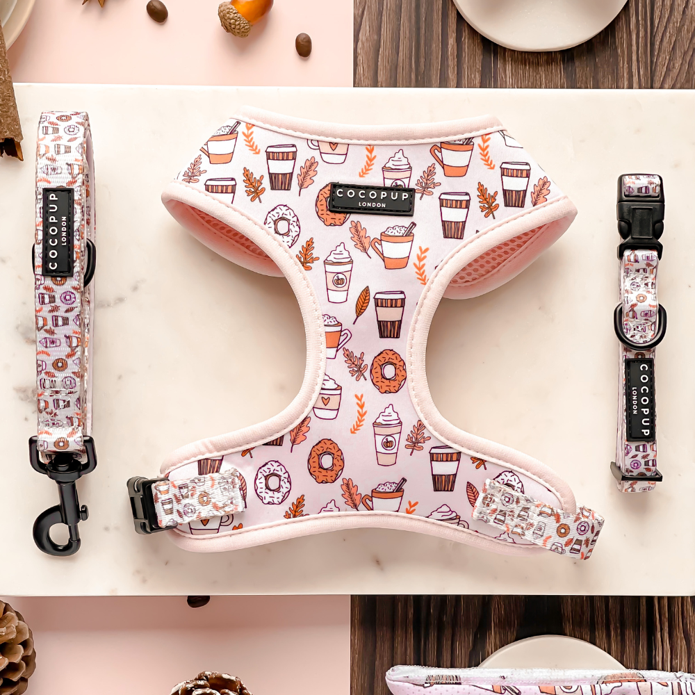 An Autumn Puppuccino Adjustable Neck Harness, Lead & Collar Bundle by Cocopup London, featuring a pink dog harness adorned with coffee and pastry designs. This set includes an adjustable harness, a matching leash, and a collar with a dog safety lock. It's displayed on a white marble surface decorated with coffee beans and pastries.