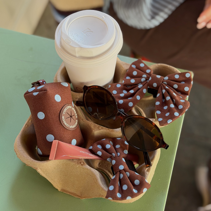 A set of brown polka-dot accessories, including a zippered pouch, sunglasses, and fabric dog bows, arranged in a brown paper coffee cup holder next to a takeaway coffee cup on a green table.