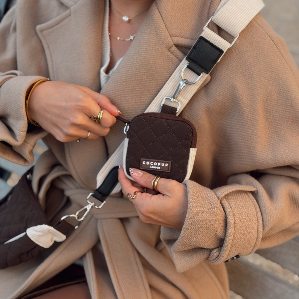 A close-up lifestyle shot of a person wearing a beige coat and holding a small quilted brown crossbody pouch with a white strap. The pouch features a silver-tone metal clasp and a brand label that reads 'Cocopup London'.