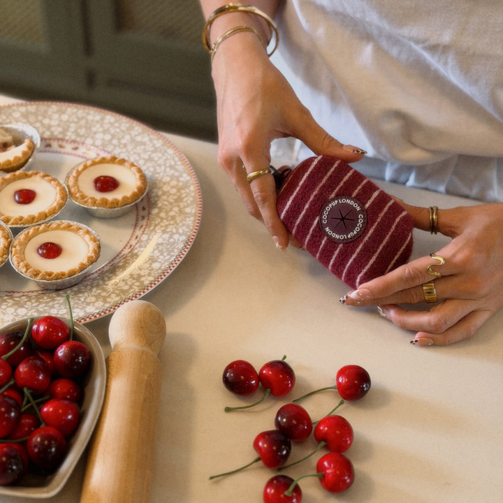 A person holds a small striped fabric poop bag holder with a circular embroidered logo on a beige tabletop. The scene includes cherry pies on a plate, cherries in a bowl, and a wooden rolling pin, indicating a kitchen or bakery setting.