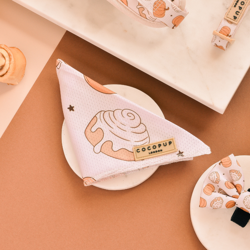 A white ceramic plate displays a triangular dog bandana featuring a cinnamon buns print and branded with the "Cocopup London" label. Nearby, matching Cocopup London bandanas with similar cinnamon roll designs are set against a brown and white background.