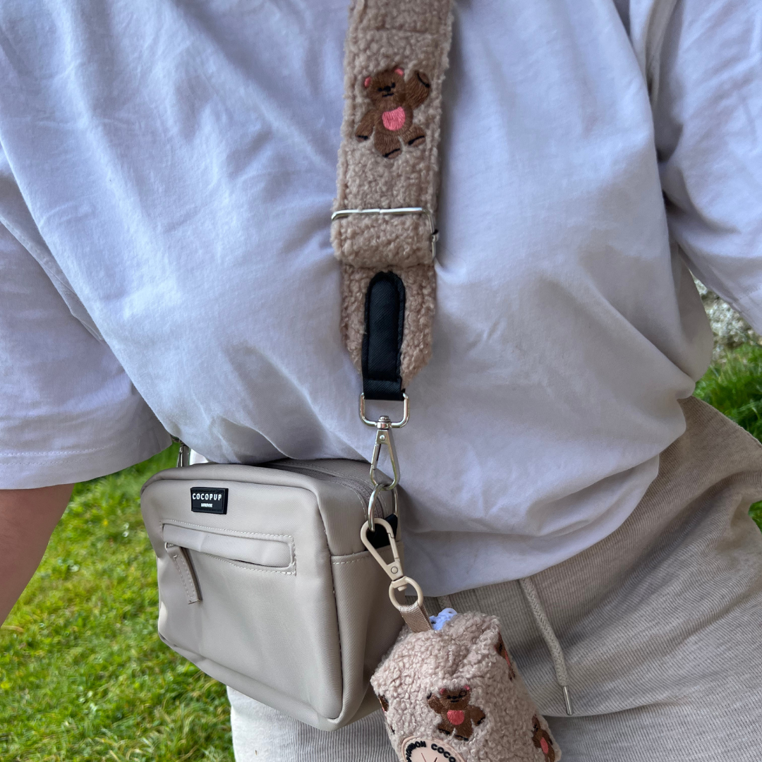 A person in a casual white t-shirt and beige pants is seen with a small beige crossbody bag. The bag strap, the Theo Embroidered Teddy by Cocopup London, features an adorable plush teddy design with a small bear holding a heart. Accompanying the bag is a matching bear-themed pouch and a small metal heart accessory, making it ideal as a dog walking bag.