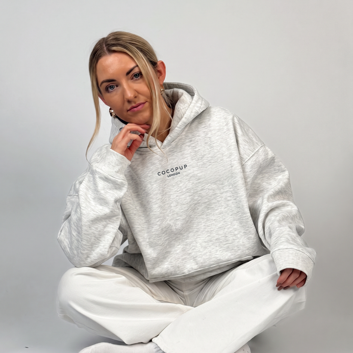 A woman wearing a light heather gray Cocodrop hoodie and white sweatpants, sitting cross-legged against a plain light gray studio background.