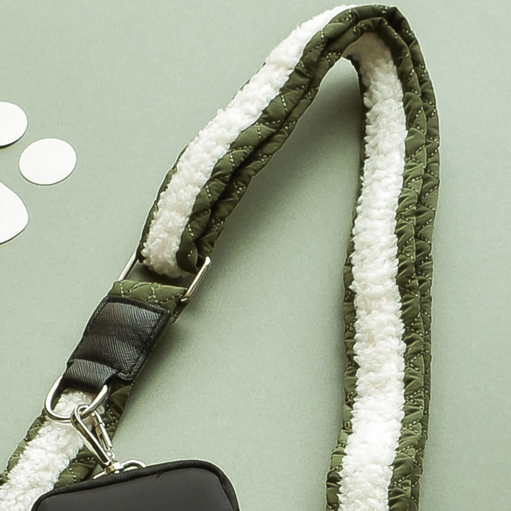 A close-up of the Cocopup London Bag Strap - Khaki Quilted with a white, soft, fur-like lining on the underside. The strap is attached to a black handbag using a silver clip. The background features a light green surface adorned with three small white circular objects.