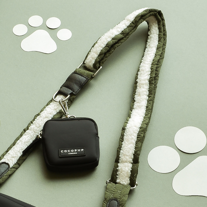 A small black pouch labeled "Cocopup London" is attached to a Bag Strap - Khaki Quilted with a white fleece lining. The Cocopup London dog walking bag items are laid on a sage green background, decorated with white paw print cutouts.