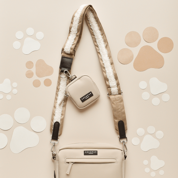 A beige crossbody bag with a detachable Nude Aviator Bag Strap from Cocopup London and a small square pouch attached is laid on a beige background decorated with various sizes of paw-print cutouts in white and brown shades. The Cocopup Dog Walking Bag and pouch feature a small black logo tab.