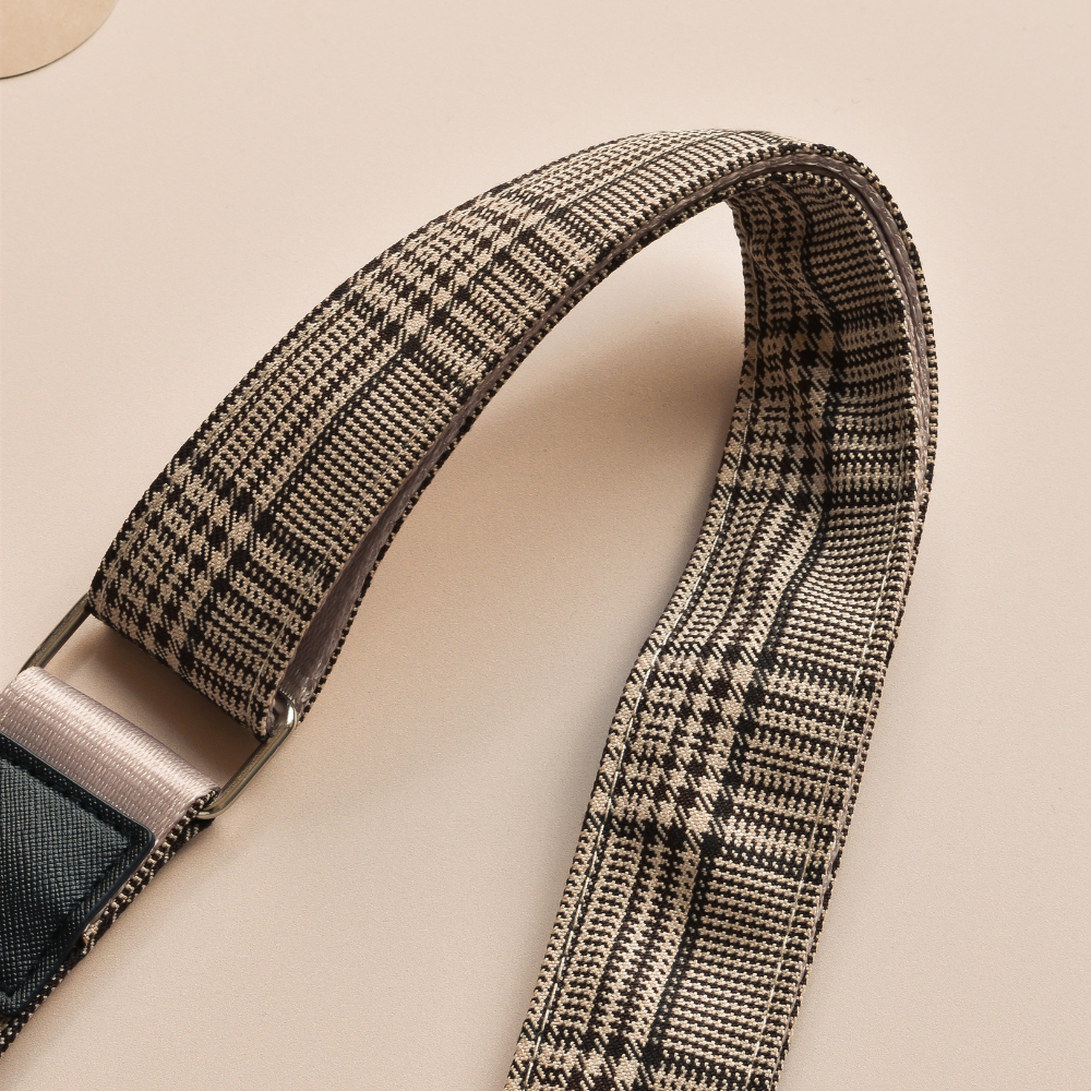 A close-up of a beige and black Bag Strap - Pup Plaid by Cocopup London, featuring metal hardware, displayed on a neutral-toned background. The strap is partially coiled, highlighting its texture and pattern.