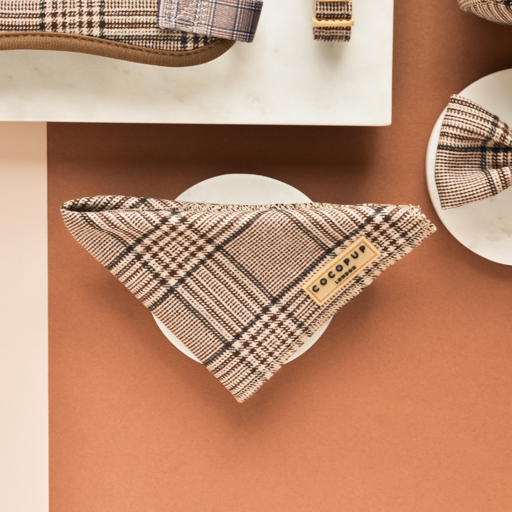 A triangular plaid Pup Plaid Bandana by Cocopup London, featuring a leather label that reads "COLLAR," is displayed on a circular white plate. The background showcases a two-tone surface in brown and cream colors, with additional plaid dog accessories partially visible in the corners.