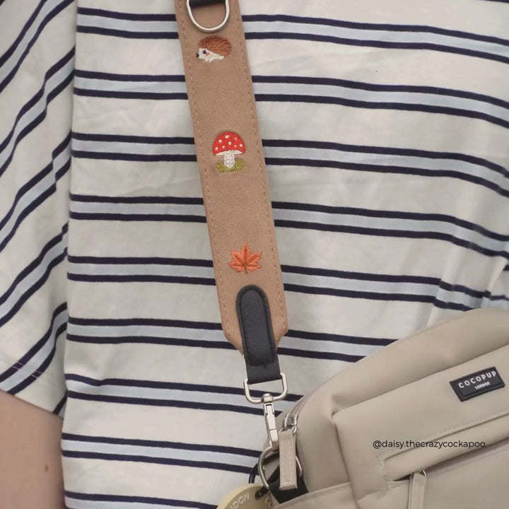 Close-up of a crossbody bag with a tan suede strap featuring embroidered red and orange mushrooms and a leaf