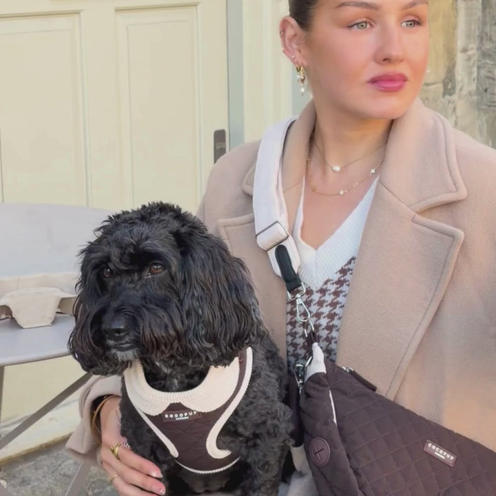 A person in a beige coat carries a brown quilted crescent-shaped crossbody bag with a strap, sitting on a bench next to a small black dog wearing a matching bag.