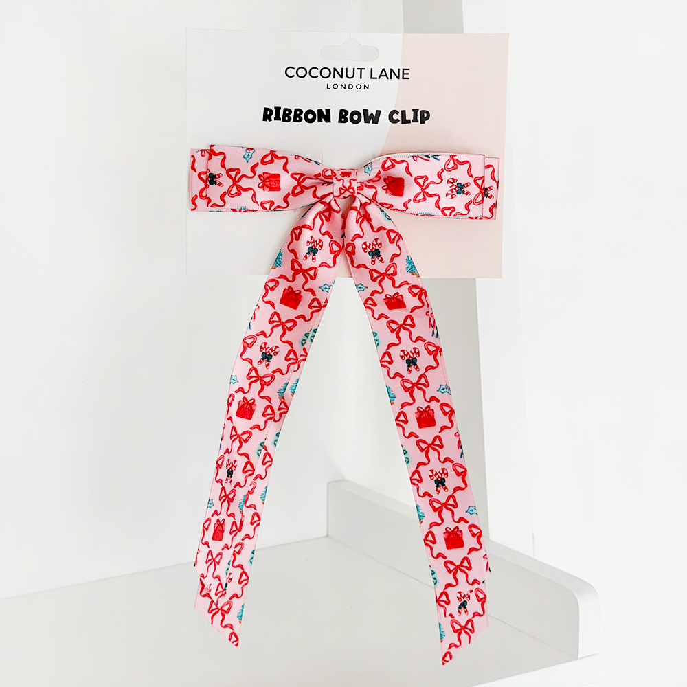 The Pupmas Kisses Ribbon Bow Clip - By Coconut Lane showcases a charming pattern of hearts and flowers, elegantly presented on a white cardboard backing labeled "Coconup London." Part of their exquisite hair accessories collection, the bow's long tails gracefully drape down the holder.