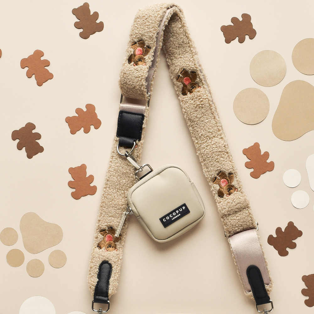 A cream-colored fuzzy camera strap from Cocopup London, known as the "Bag Strap - Theo Embroidered Teddy," featuring teddy bear face designs, is laid flat on a beige surface. Attached to the strap is a small, square, beige Dog Walking Bag with a black "ECO COUP" label. Various brown and beige paper cutouts in different shapes are placed around the strap.