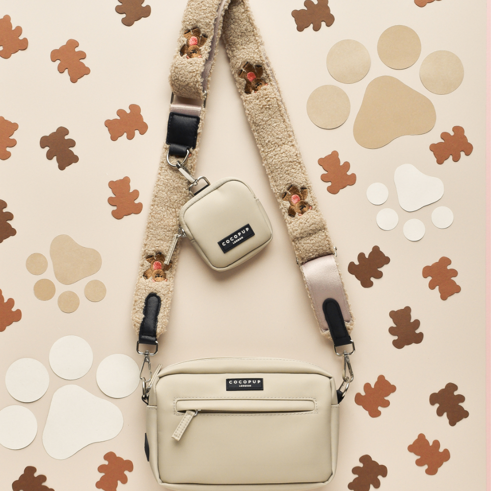 A beige crossbody dog walking bag with a matching mini pouch is attached to the Theo Embroidered Teddy strap by Cocopup London. The bags are displayed against a background adorned with various brown, beige, and white paw prints and bear shapes.