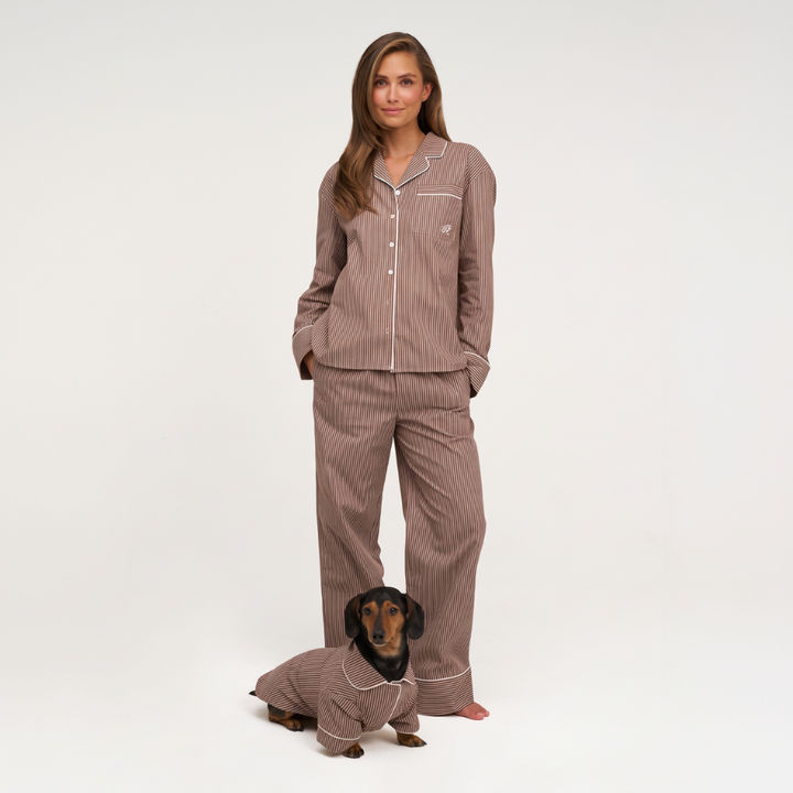 A woman wearing a brown pinstriped pajama set stands barefoot against a white studio background, with a small dachshund dog sitting in front of her wearing a matching pajama top. The pajamas have a warm taupe-brown color with thin white pinstripes and white piping.