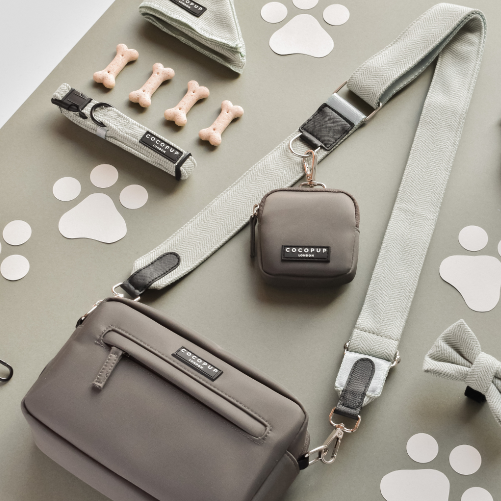 A flat lay showcases the Dog Walking Mocha Bag Bundle - Sage Tweed by Cocopup London, featuring various dog accessories in neutral tones, including a crossbody bag, treat pouch, bandana, bow tie, leash, and collar. Additionally, there are small dog bone-shaped treats and paw print cutouts scattered around.